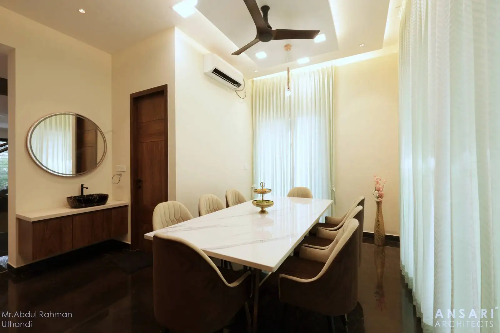 The dining room showcases a sleek marble-topped table for eight, complemented by floor-to-ceiling windows with sheer curtains for natural light. A stylish console with a vessel sink and mirror adds luxury and functionality, making the space ideal for dining and entertaining.