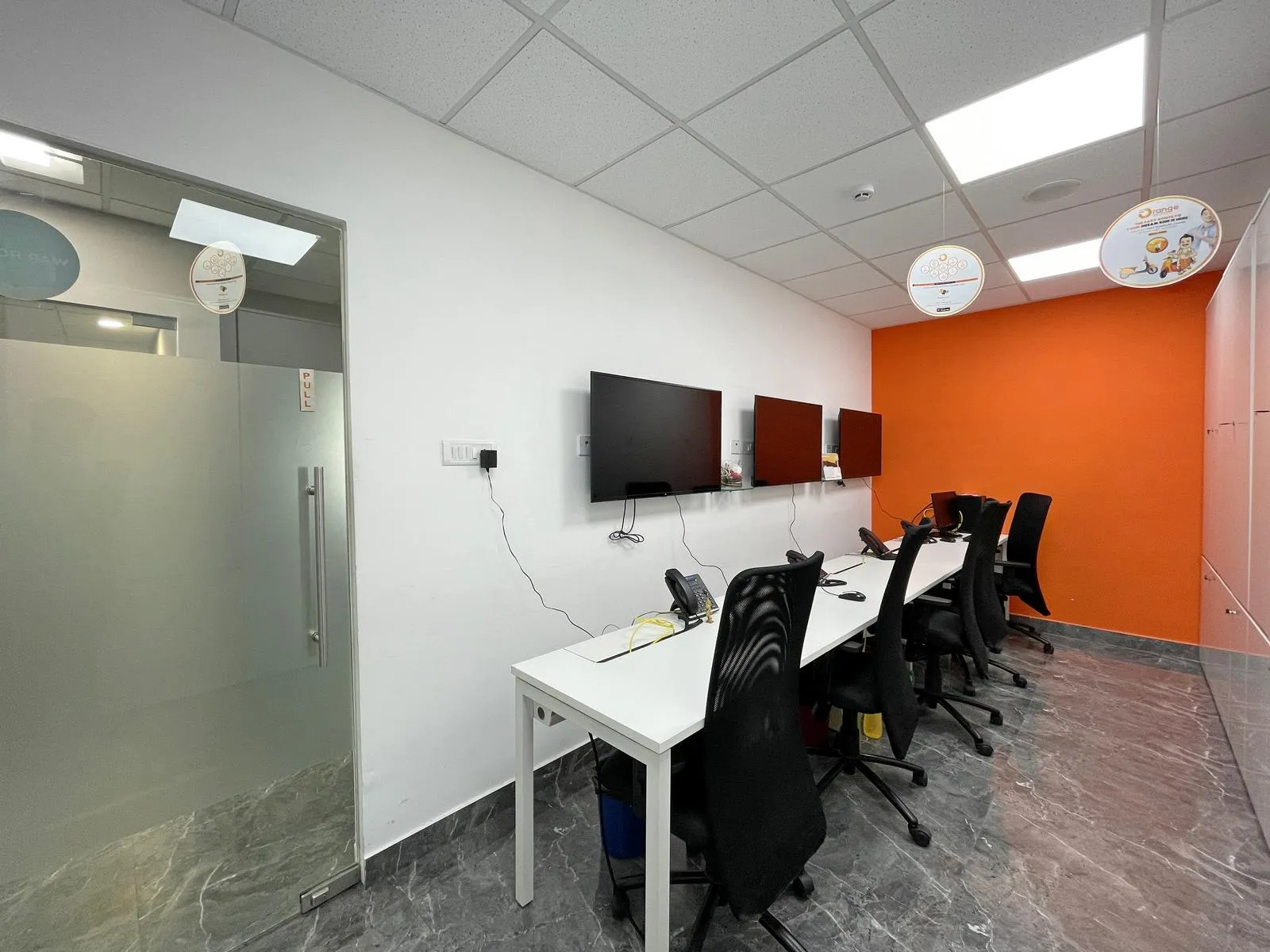 The 5-seater war room combines ergonomic workstations, wall-mounted displays, and an orange accent wall for a modern, productive, and professional workspace.