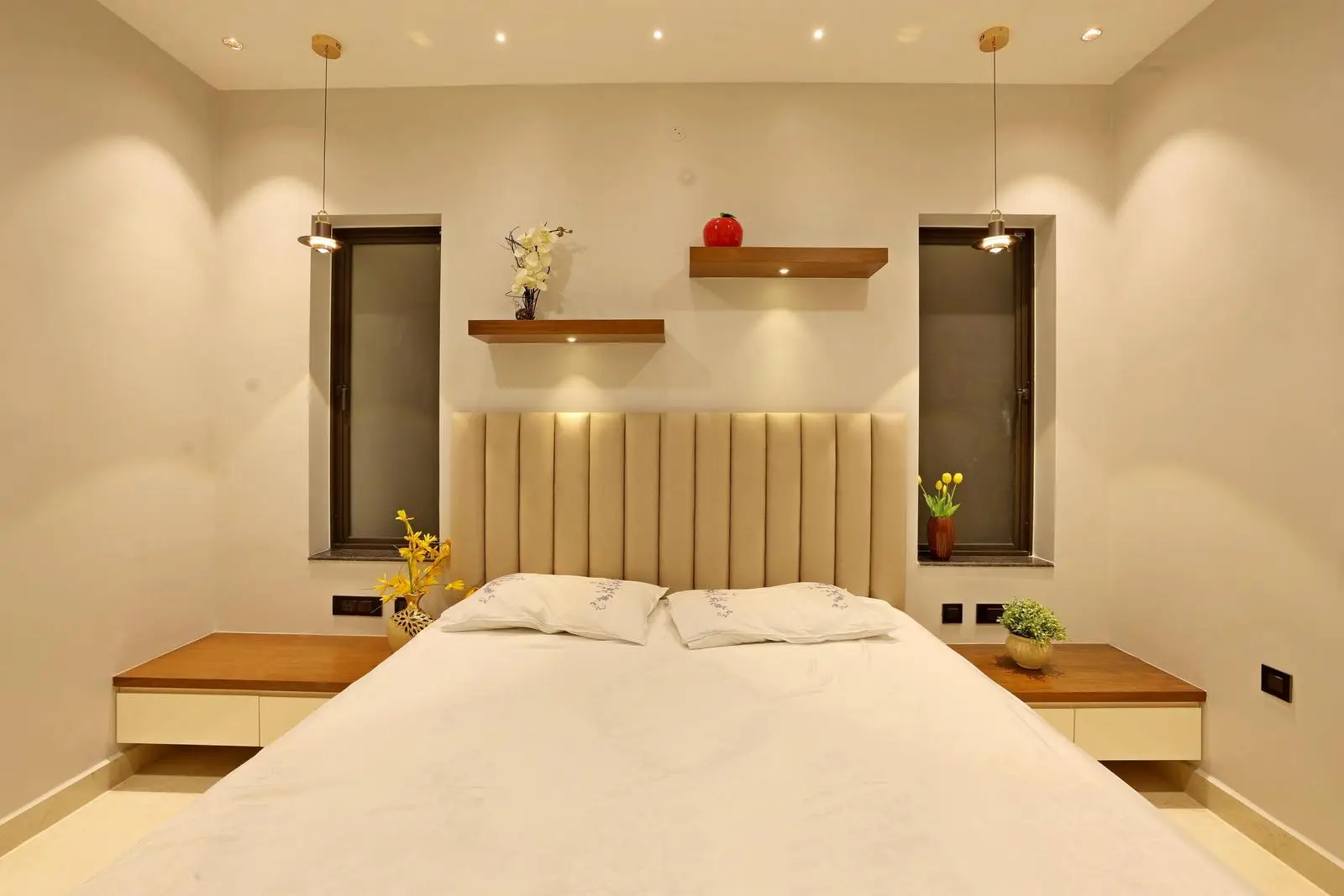 The guest bedroom features a centered bed with vertical windows and floating nightstands. Beige tones, wood accents, and an upholstered headboard create warmth, while recessed and pendant lighting enhance the minimalist, inviting design.