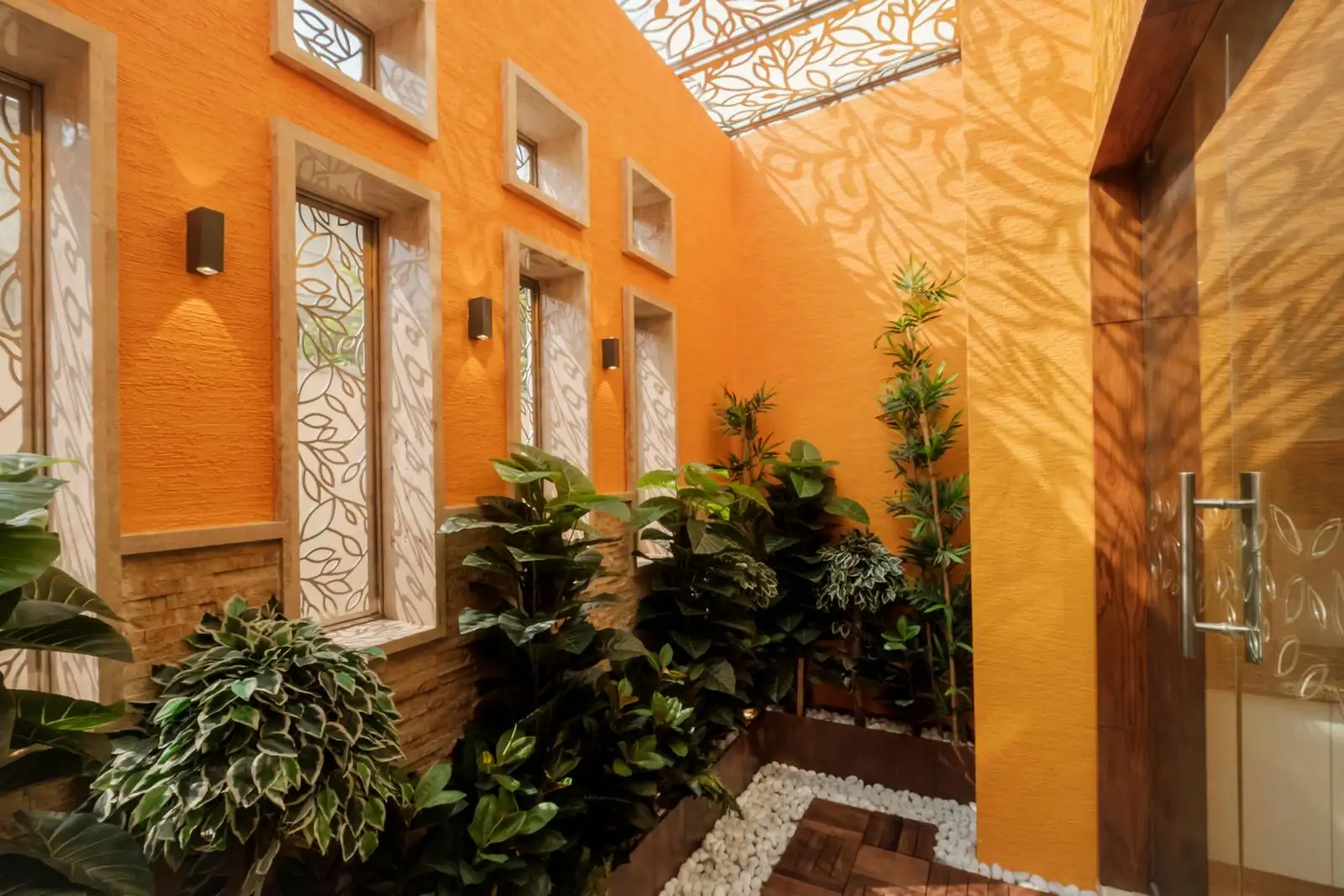 This garden space features a harmonious blend of texture, light, and greenery. The orange textured walls and narrow frosted-glass windows with a leaf motif create a vibrant yet serene backdrop.