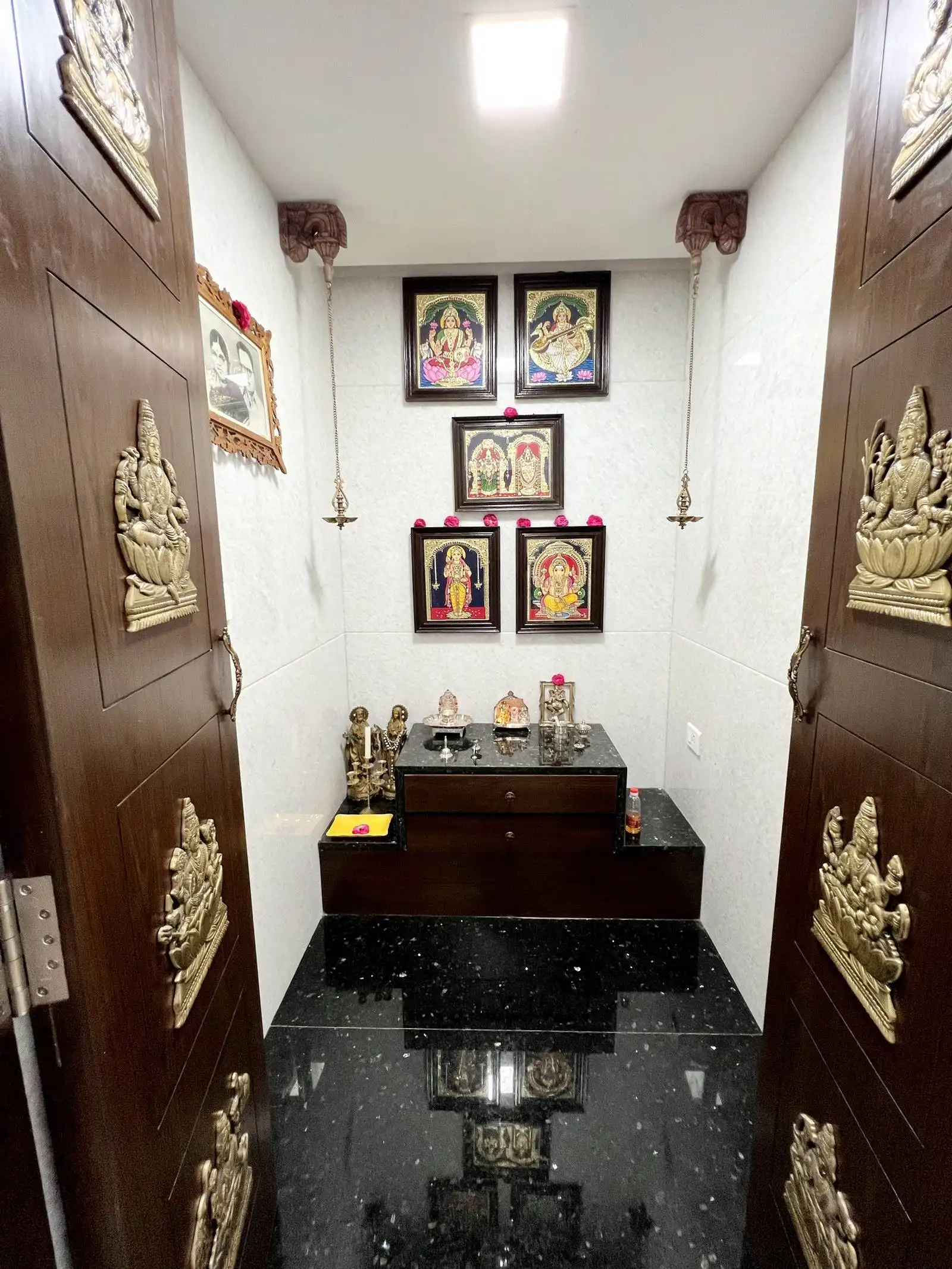A serene pooja room with polished black granite floors, white tiled walls, a traditional wooden altar, and warm brass accents for a peaceful prayer ambiance.