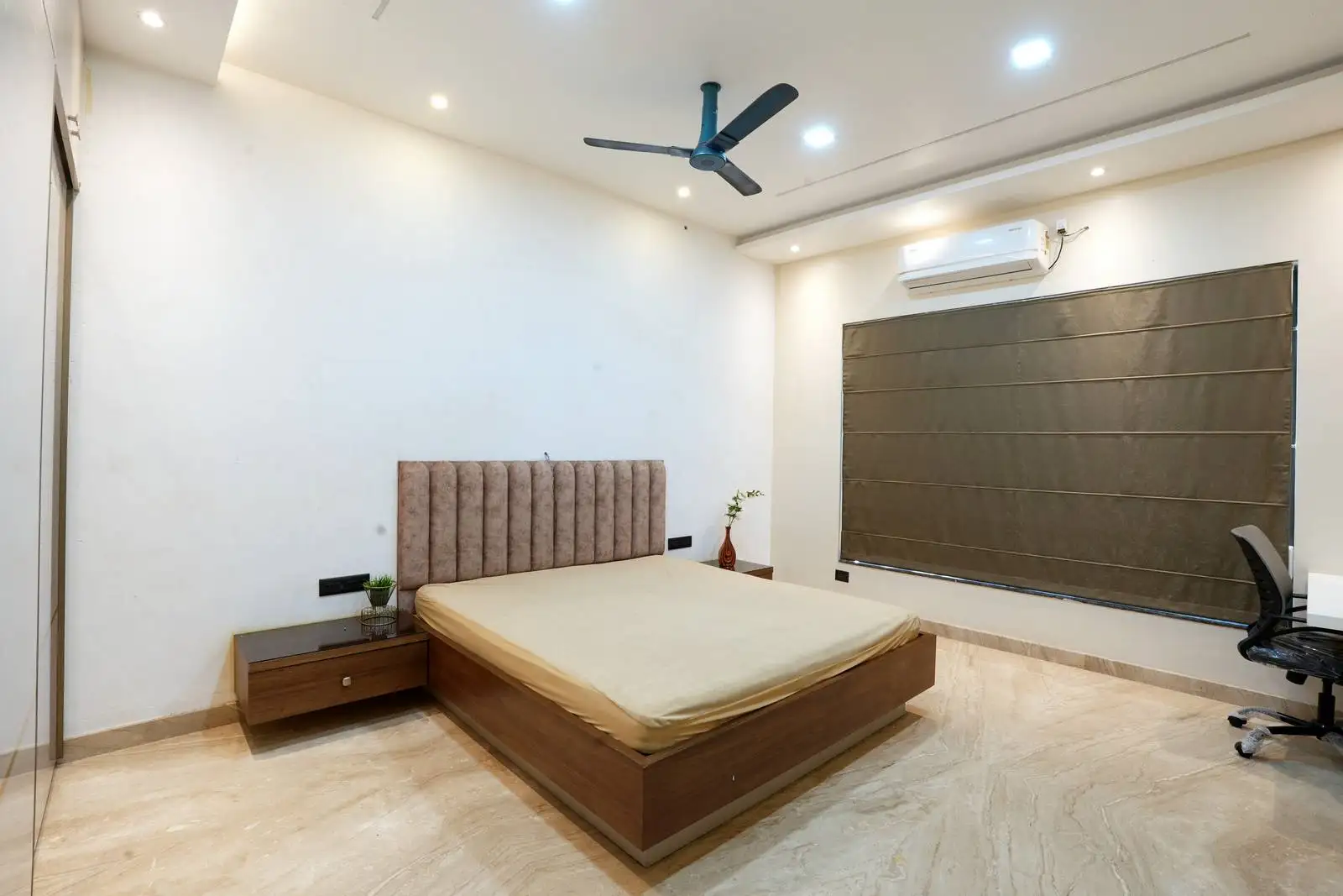 Warm wooden bed with beige tones, minimalist nightstands, and recessed lighting create a calm atmosphere. Large window with dark curtains and light flooring enhances space and comfort.