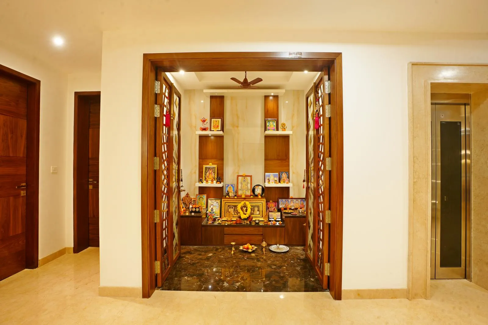 The puja area ( 6'0" x 8'0" sq. ft ) is recessed into a niche with an elevated deity platform, wooden shelves, and warm lighting, creating a serene and sacred space. Dark marble flooring, light beige walls, and wooden accents enhance the spiritual ambiance and visual focus.