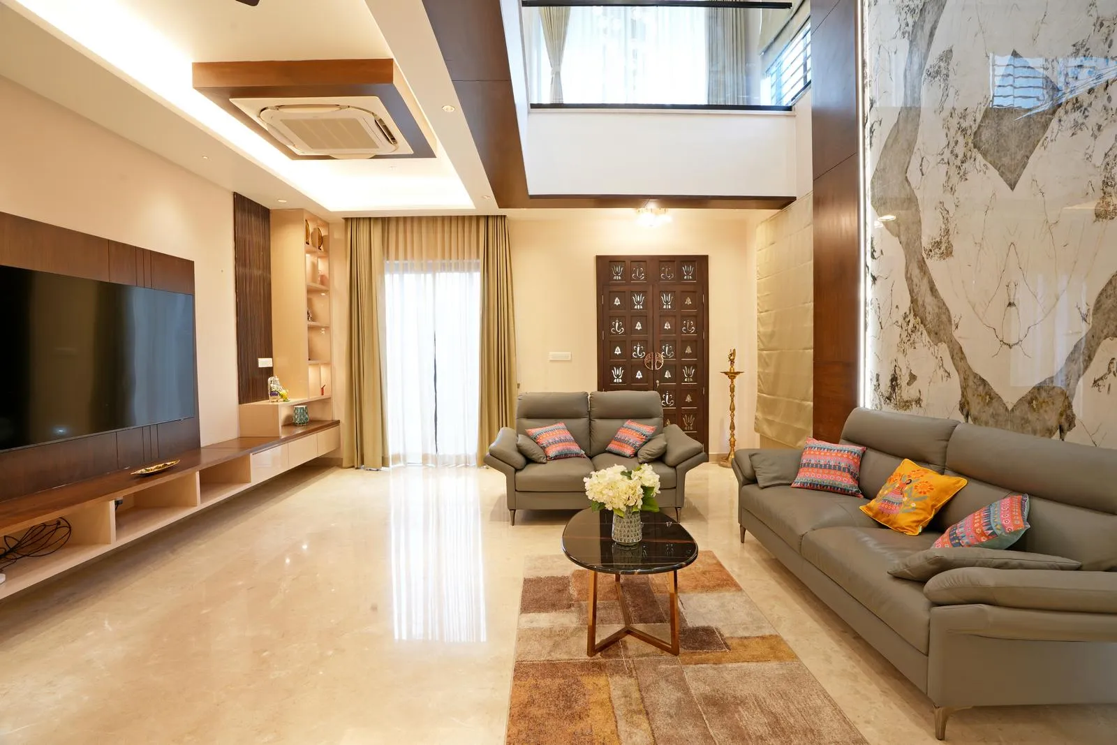 The first-floor living area ( 17'0" x 18'0" sq.ft ) features a double-height volume with polished marble flooring, a feature TV wall, and wooden accents for material harmony. Open-plan layout, balcony connection, puja room partition, large glass doors, and recessed lighting enhance vertical openness, natural light, and seamless spatial flow.