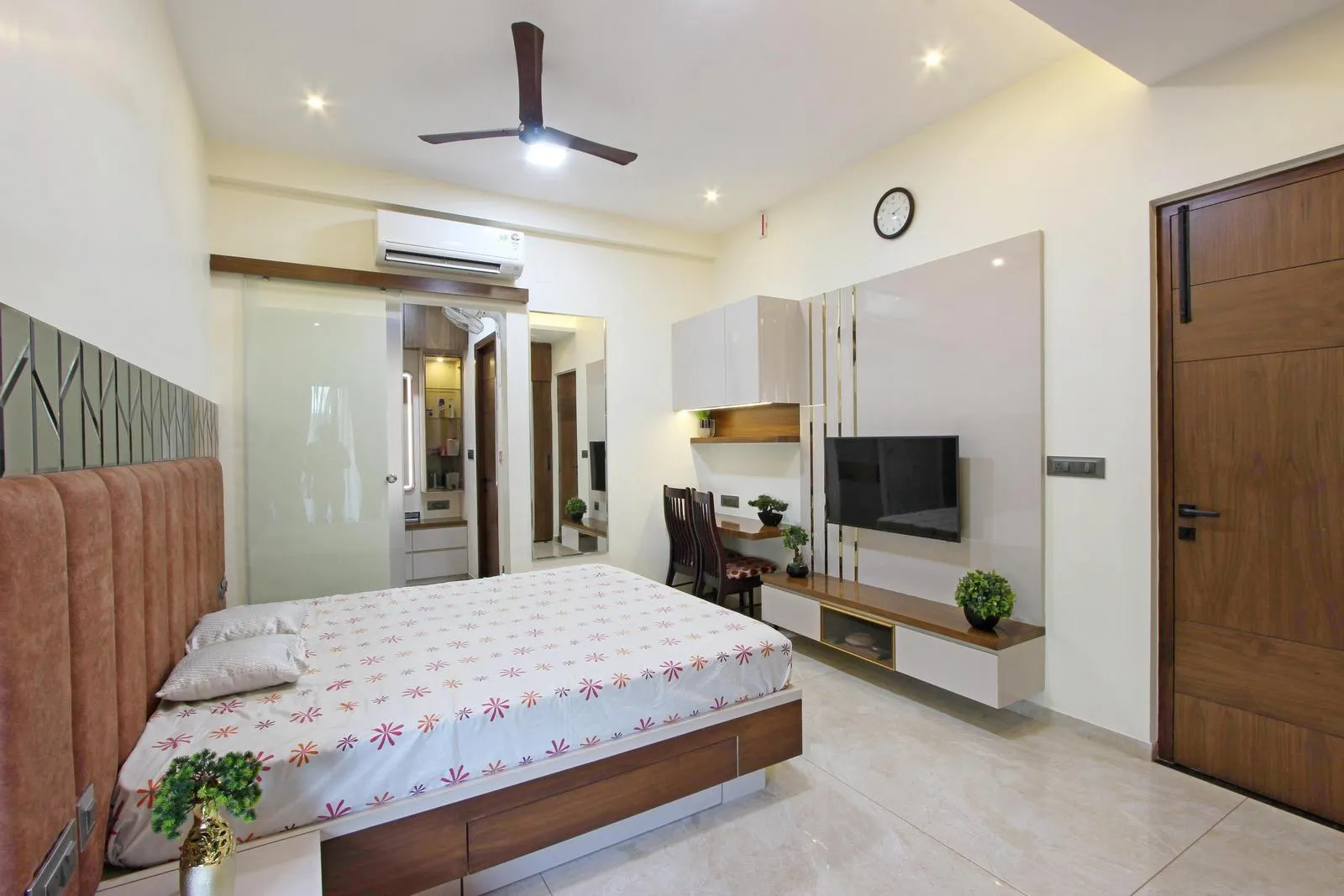 This bedroom has a central bed, an integrated TV unit with study desk, and a frosted glass bathroom door. Warm walnut tones, light flooring, and a modern palette create a bright, functional, and cozy space. The rectilinear layout and reflective surfaces enhance circulation, depth, and visual flow.