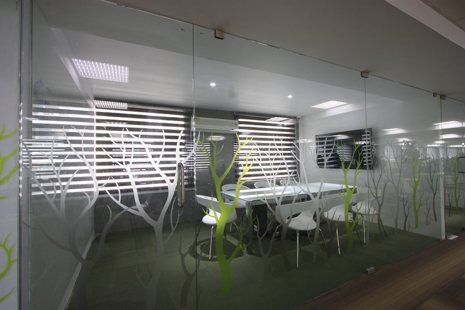 Glass-enclosed meeting room with frosted green tree patterns, white furniture on a green carpet, and a wall-mounted screen for presentations.