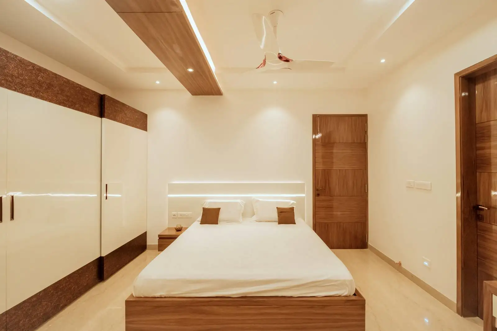 Ground floor bedroom features polished light marble flooring for brightness and luxury, paired with warm wood-toned bed frame, wardrobe, doors, and ceiling panel. Glossy white and wood-accented wardrobe blends modern and natural aesthetics.