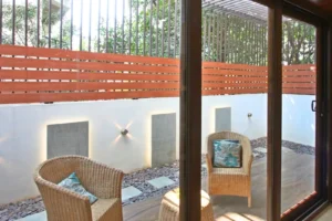 Small outdoor patio with two wicker chairs each with a blue patterned cushion, a matching wicker side table, surrounded by a white wall with embedded lighting and horizontal wooden slats on top making a fence, viewed through glass sliding doors.