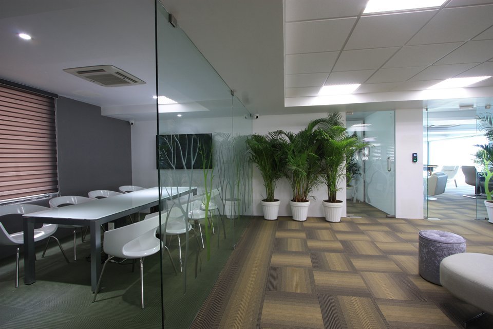 Office interior featuring a glass-walled conference room, white table and chairs, potted plants, tufted stools, and patterned carpet tiles under bright ceiling lighting.