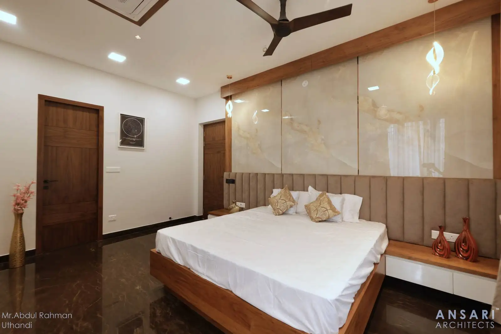 The bedroom is spacious, with a modern, minimalist design. The bed is centrally placed against a feature wall with a soft, upholstered headboard and integrated side tables.