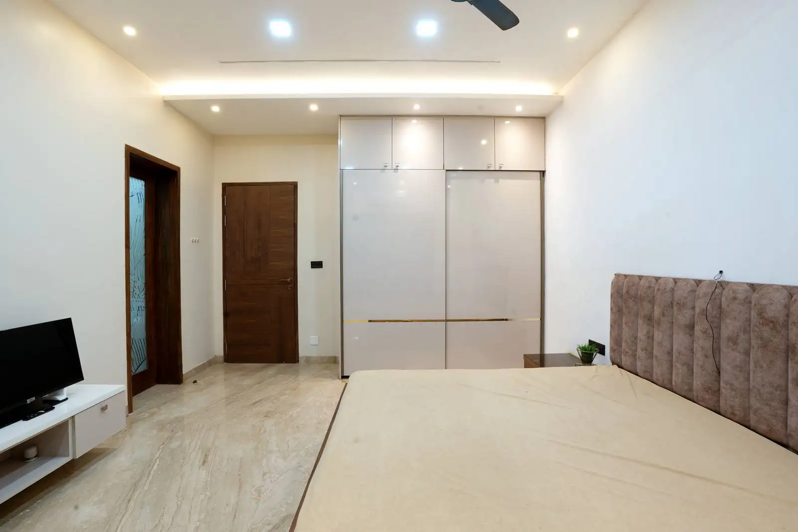 Elegant bedroom with a tufted beige bed, glossy white wardrobe, and marble flooring.