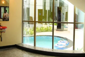 Indoor view through large glass windows showing a round pool with blue water and two colorful inflatable rings floating, surrounded by a tiled edge and plants, with a beige wall and red sink on the left side and a reflection of a man in a mirror.