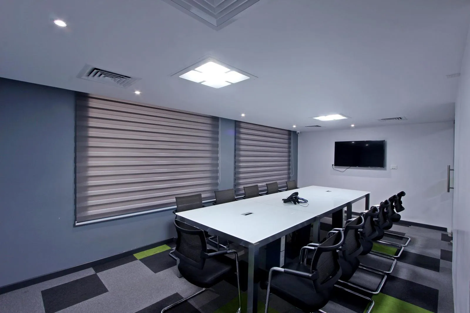 The conference room features a white rectangular table with ergonomic chairs, modular carpet flooring, and dual-tone walls, creating a professional and visually engaging environment. Integrated LED lighting, air-conditioning, and a wall-mounted display enhance comfort, functionality, and digital collaboration.