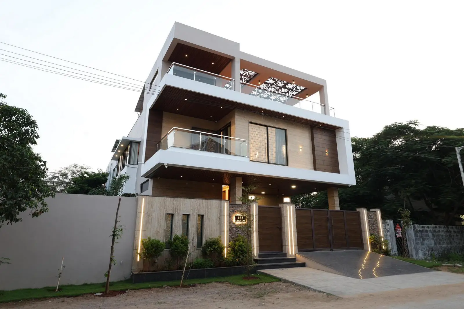 The main gate blends wood and metal, while glass balcony railings ensure openness. A perforated terrace pergola adds shade and style, and landscaped boundary greenery softens the modern exterior.