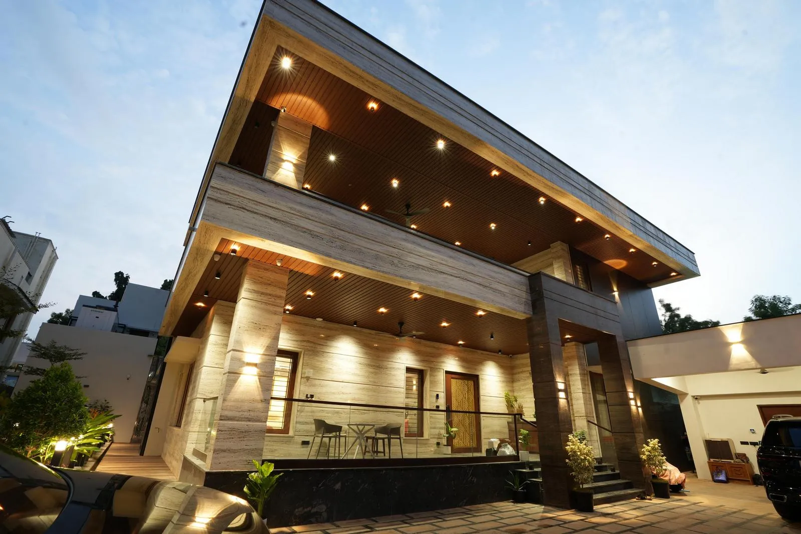 Modern two-story house with extensive use of warm wooden and stone textures, recessed lighting on ceilings and walls, glass railings on the balcony, and a tiled driveway with plants and parked cars.