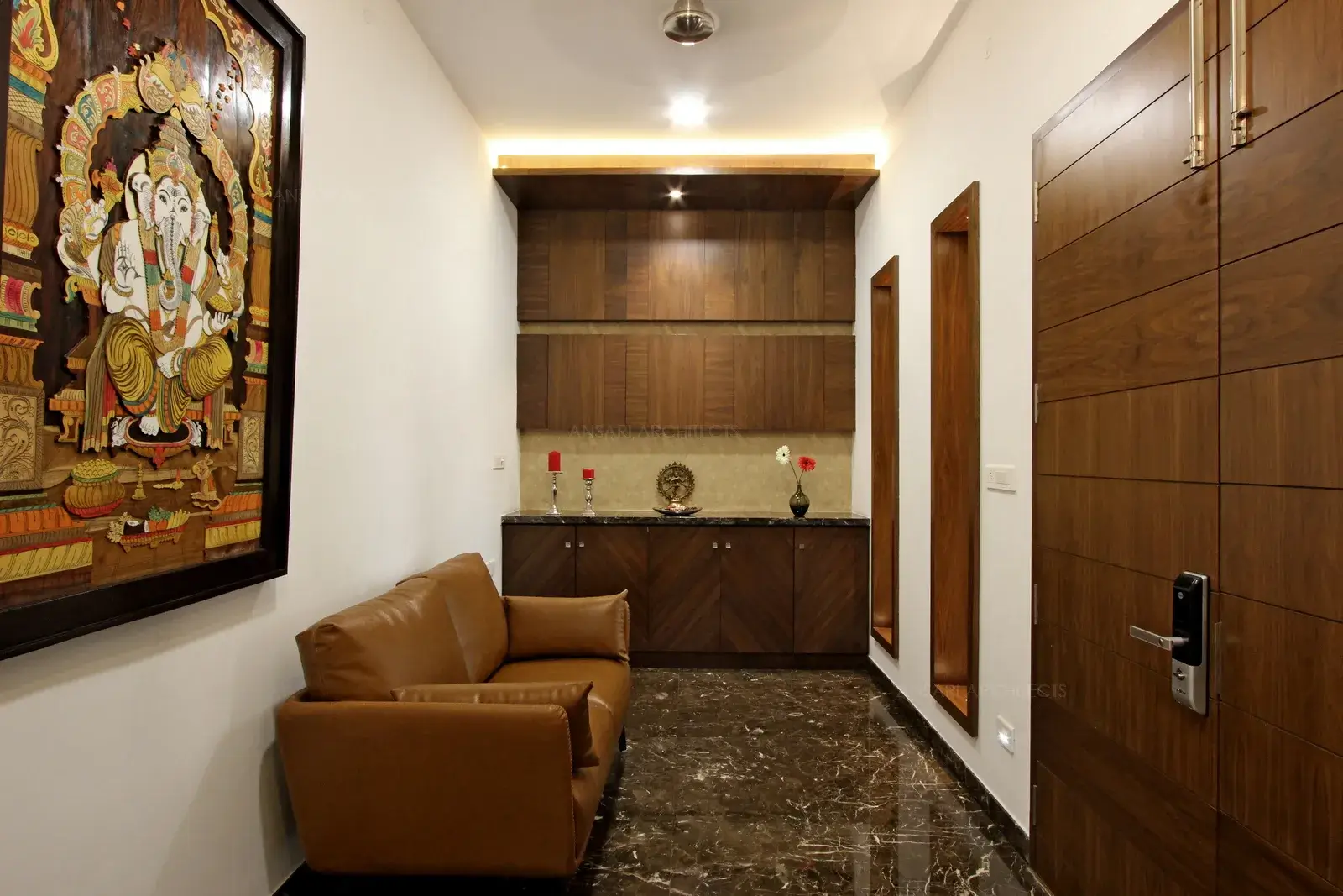 The Entrance foyer space is well adorned with wood panelling, Featuring intricate artwork and useful storage for newspapers, umbrella, etc below. The colourful painting catches the eye and sets the palatial vibe that runs through the house. Brown sofa, panel lights, and wooden textures that are highlighted with different variations create an inviting ambience.