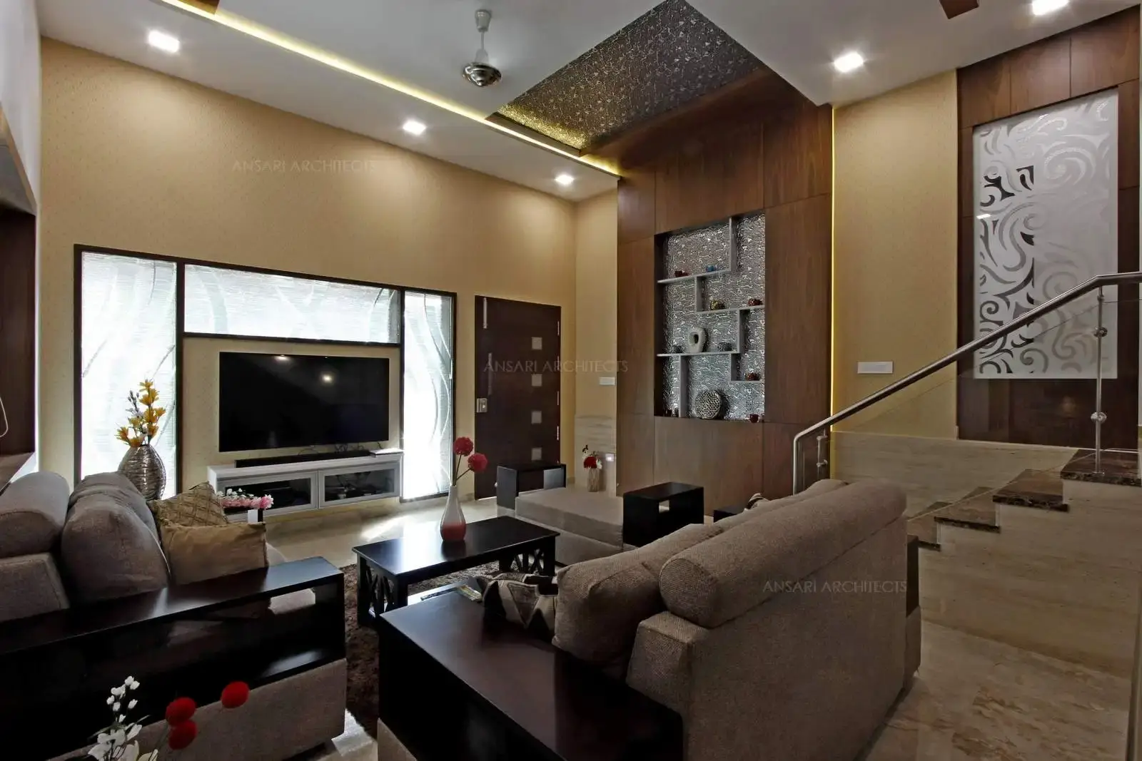 Multiple colours work together here. The monochromatic wallpaper creates a perfect backdrop to the colours in foreground. The white ceiling with spotlights and the wooden false ceiling on either sides with silver lining recessed onto it and the cove lights embellish below. The Tv unit wall is the focal point of the living space and is remarkably designed with tinted glass on three sides. Simple and sleek boxes with clean edges Tv sits on a monochrome background and highlights the arrangement.