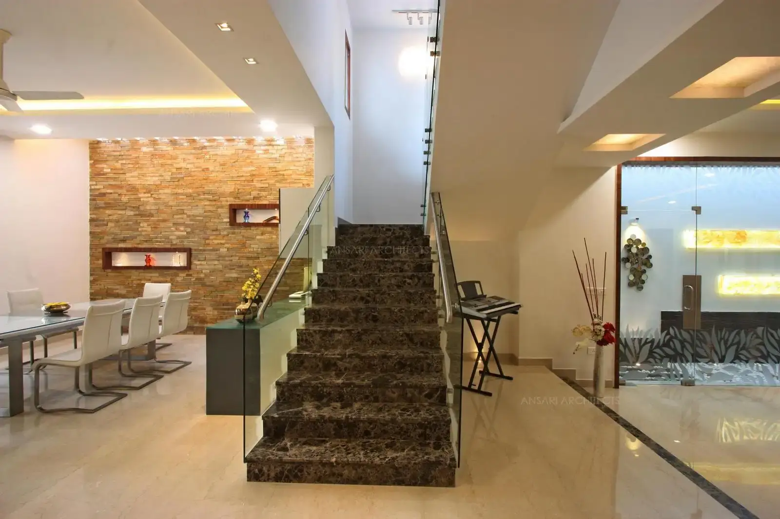 Staircase creates a strong focal point of the house. The staircase looks as emerging out of the floor in a dark toned textured brown coloured treads lending a touch of elegance to the room and warms the modern setup with glass handrails and smoothing white surrounding matches the walls.