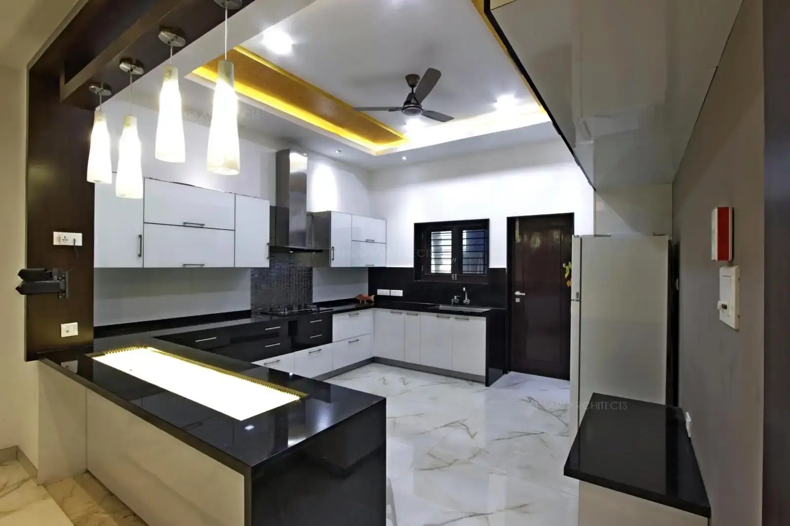 Here a row of pendants and covelights above the counter bring warmth to the crisp white setting , the counter looks light with granite top. Kichen is tactfully planned to be a decluttered space, light tones, storage cabinets at the bottom combine to give a breezy appeal. The floating shelves allow minimal storage.