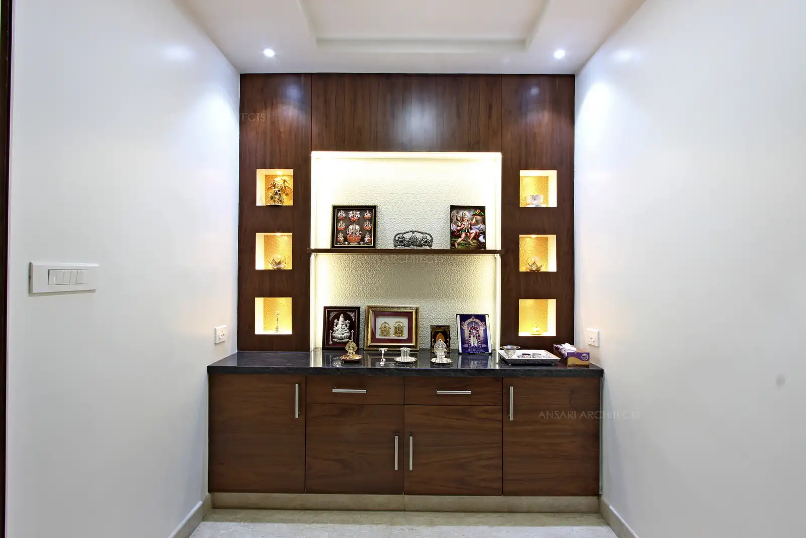 The puja room is illuminated with spotlights in a gentle glow enveloping the space in a ethereal radiance. The recessed lights in the ceiling throw down a warm glow that is complemented by the wooden ceiling and marble floor.