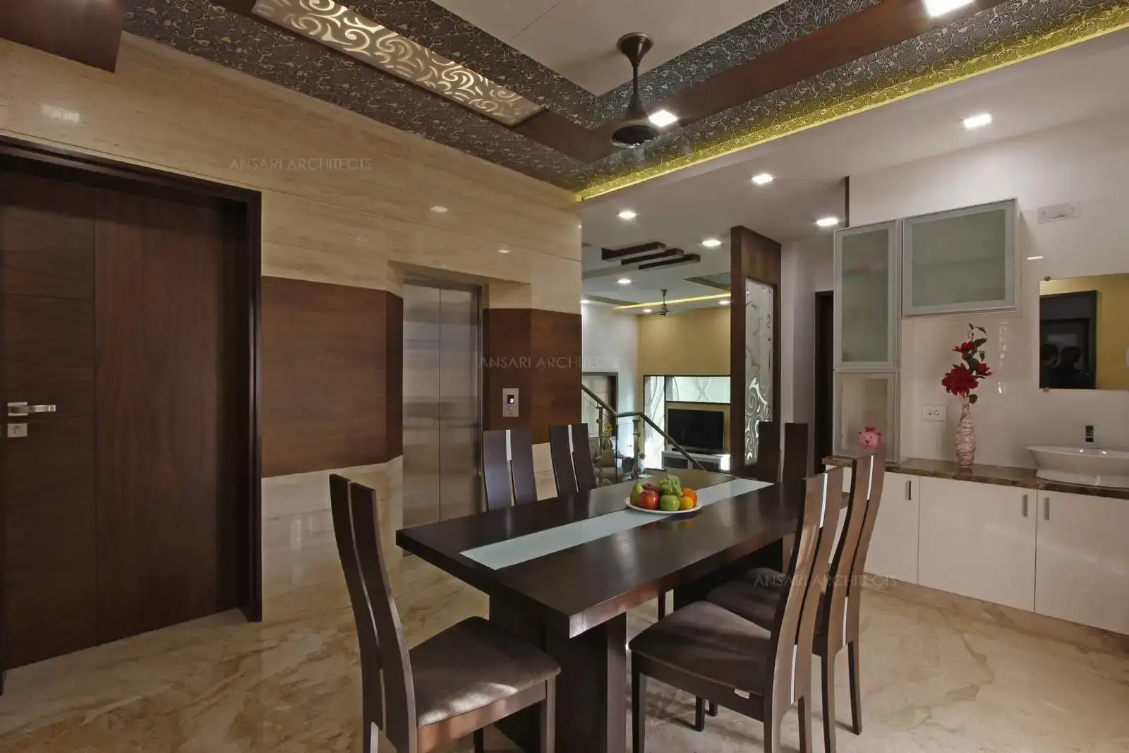 The clean lined solid wooden dining table and chairs inspires for a great formal seating and the table achieves a long rustic feel. This area is juxtaposed with the living area but set on a raised platform entirely peppered with bright spot lights and embroidered cushions. The wooden doors contribute to earthy setting. Ceiling offers two silver lined frames offset with wooden panelling and central part is projected which is perfect contrast .and the patterns on either sides of the panel giving charm to the space.