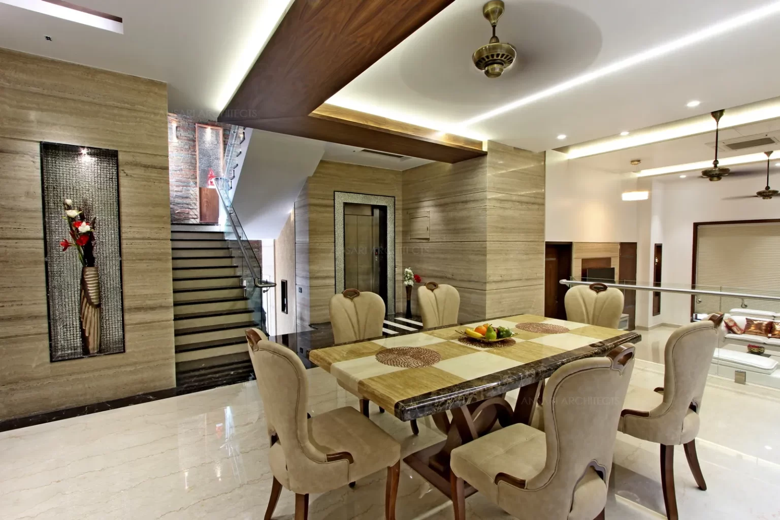 The marble clad walls with recessed niches influences the space . The dining table top has a unique chequered pattern . The veneer false ceiling infuses a grater degree of warmth.