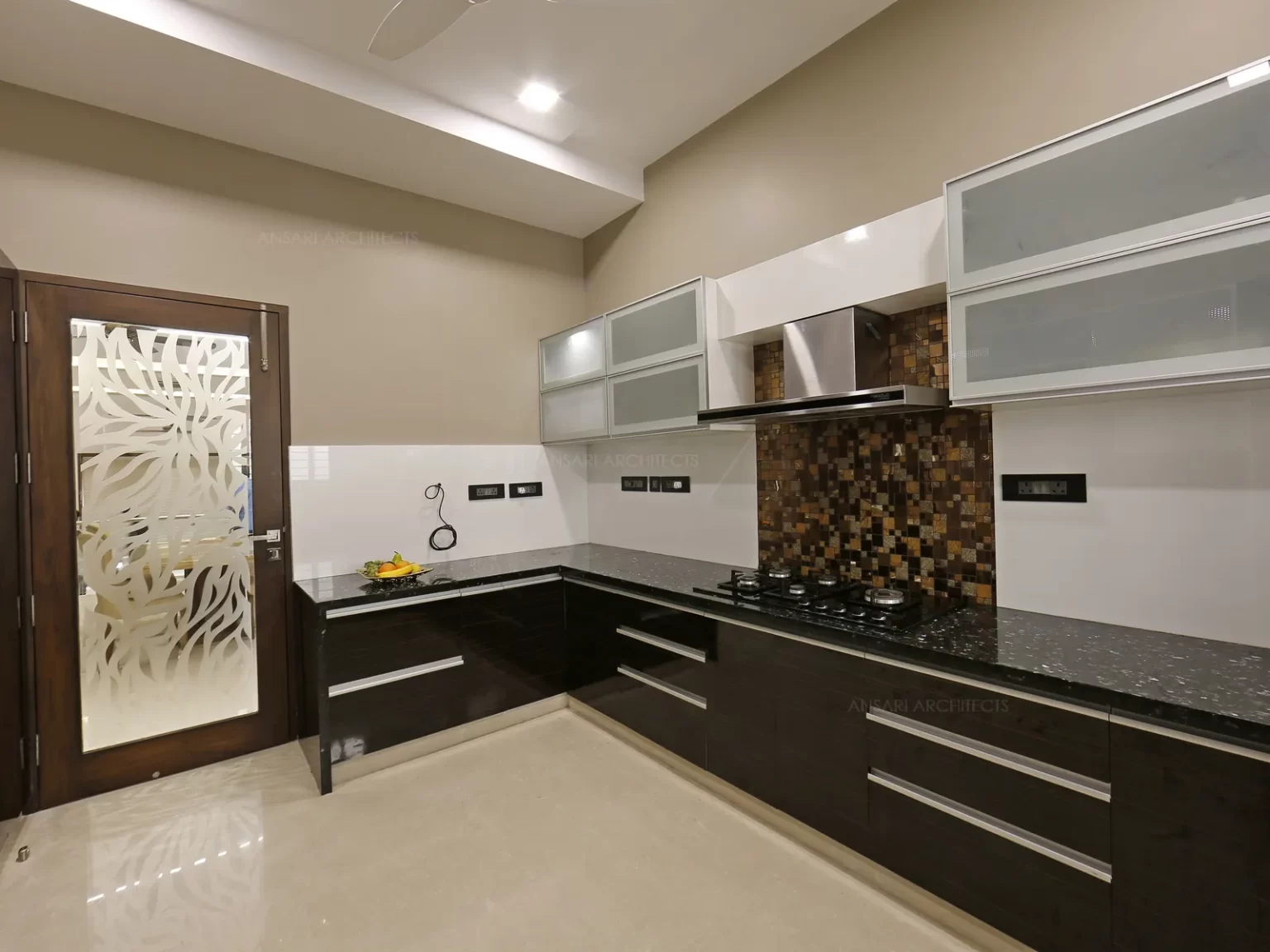 The granite kitchen top has sparkling appearance where the entire space is smart and well integrated. Spotless handle free cabinets give a clean and uncluttered look.The backsplash tiles have a vibrant and neutral hues black, brown and beige with lights under overhead cabinets adds a comprehensive nature.