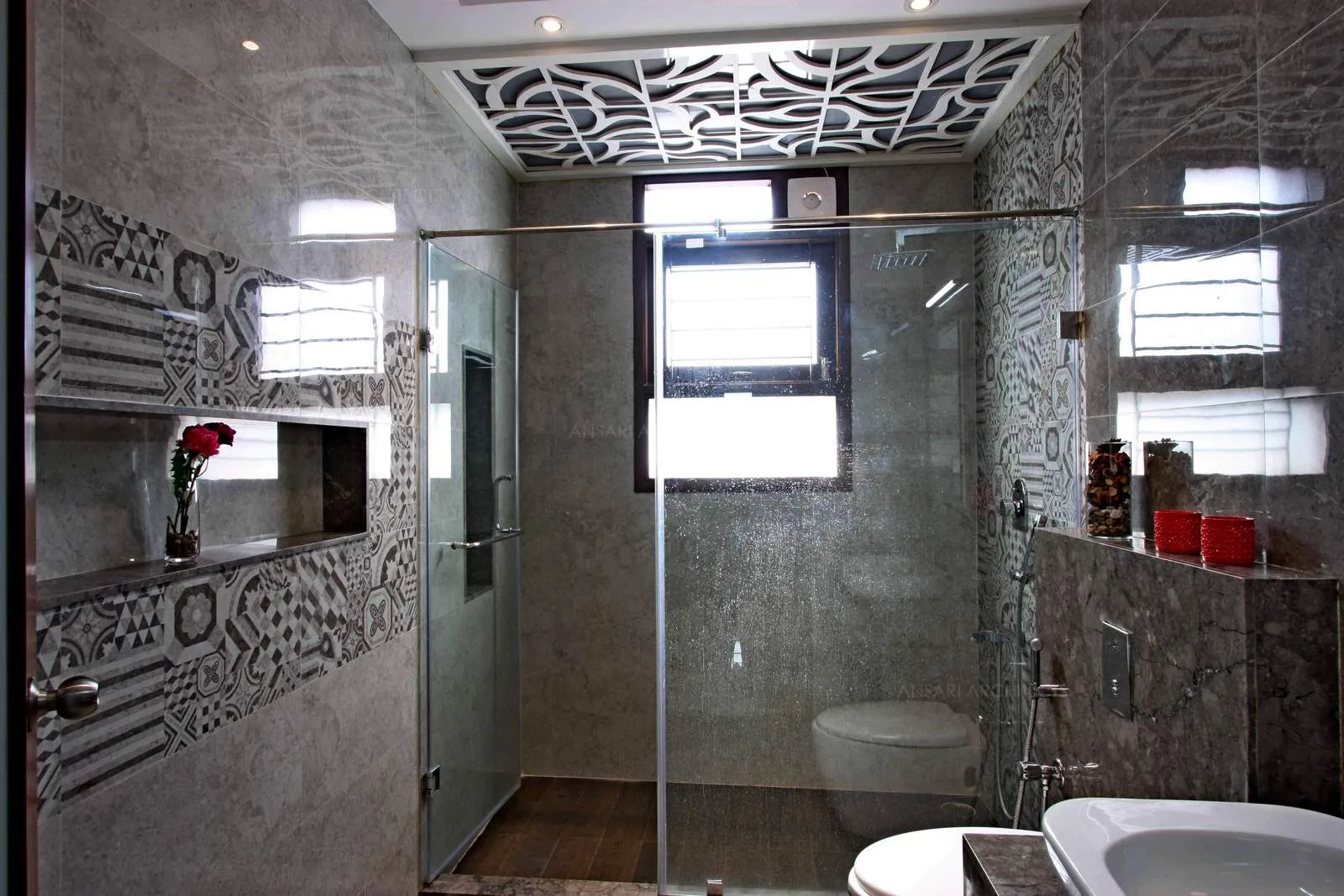 The bathroom space with vitrified tiles of specific patterns, dark tone enhance the outlook of the area with decorative panels of abstract panels, shower area is delineated with glass panels and wooden texture flooring. Niches are a special feature for the space.