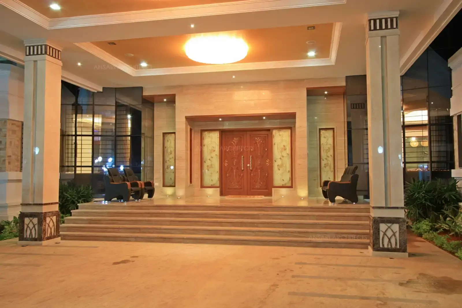 An elevated veranda with main entrance has rich wooden carved door with designer glass panels surrounded by marble cladding all around.