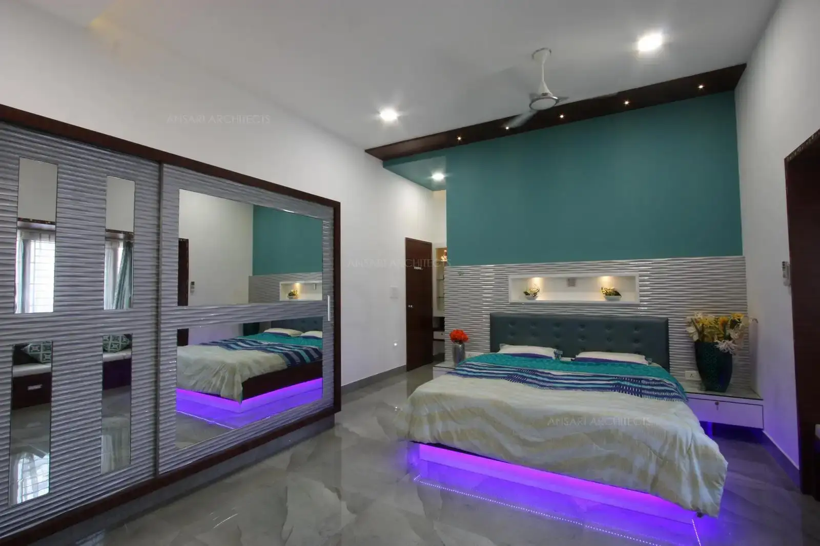 The purple backsplash light beneath the bed enhances the space, the flooring brings a sense of depth and some respite from the lushness of purple. The headboard is a textured, the cushion aptly complement the setting, niches provide a quirk with lights. The mirror ruse on the wardrobe panels makes a modish statement, this features exact texture as of the headboard and completes the space.