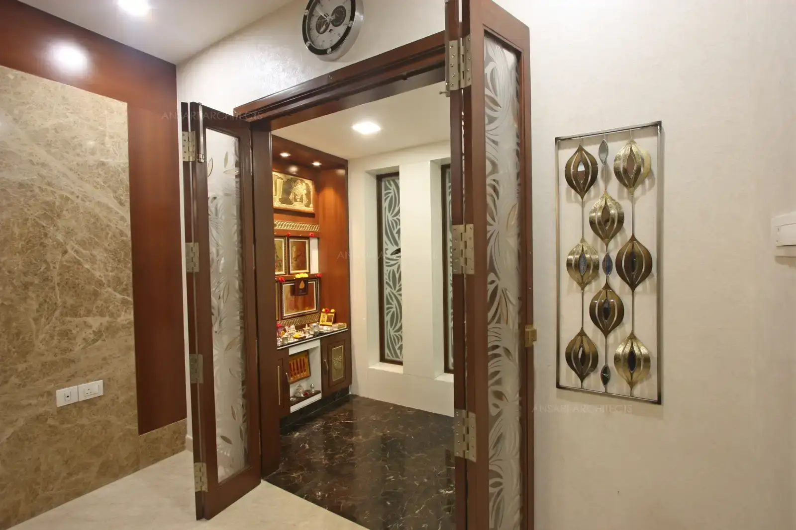 The puja room exudes warm and inviting serenity, dark flooring creates a balance with the white walls, opening with decorated stencilled motifs bordered on intricately designed panels.