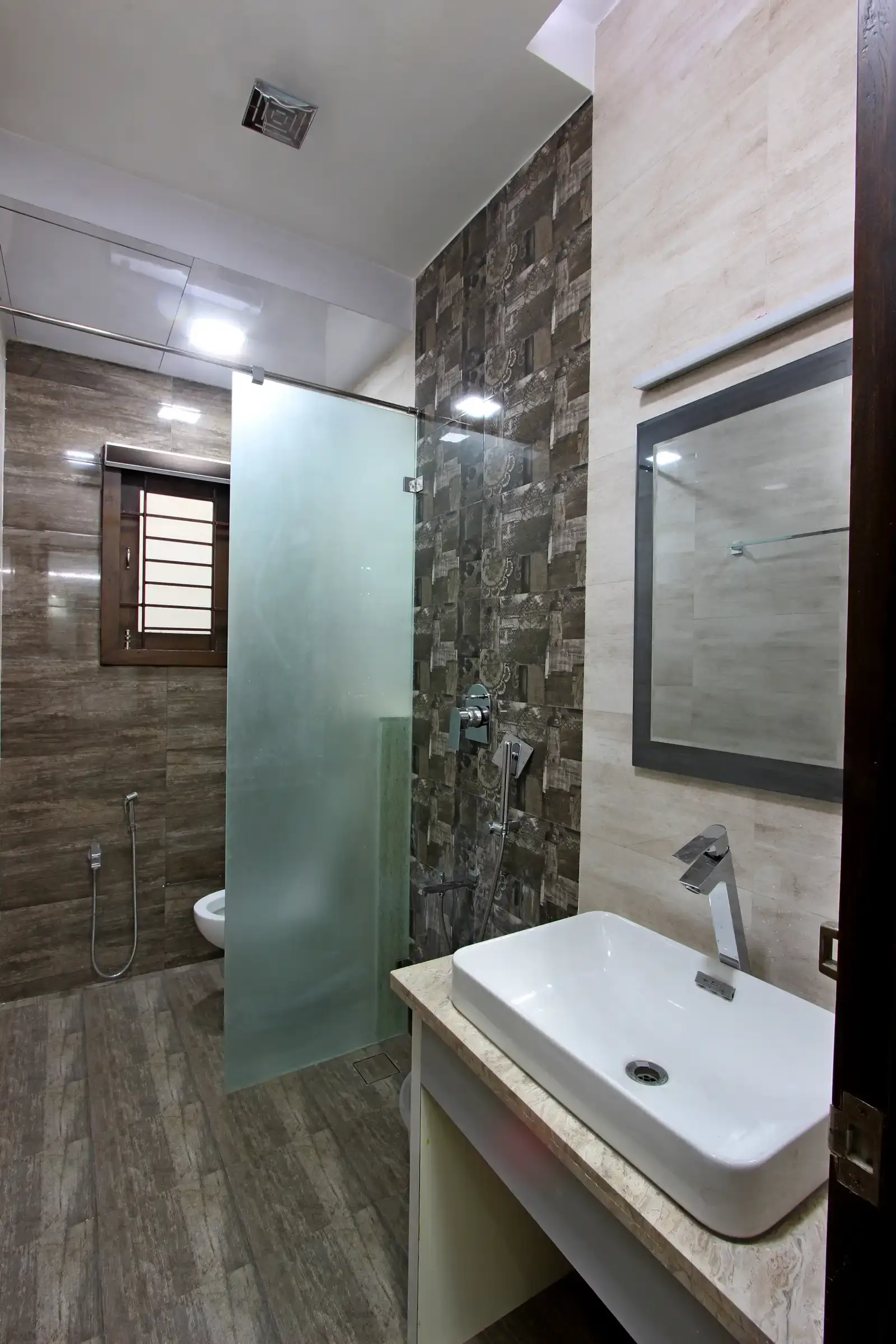 The bathroom has decorative patterned tiling on walls and dark coloured vitrified tiles .