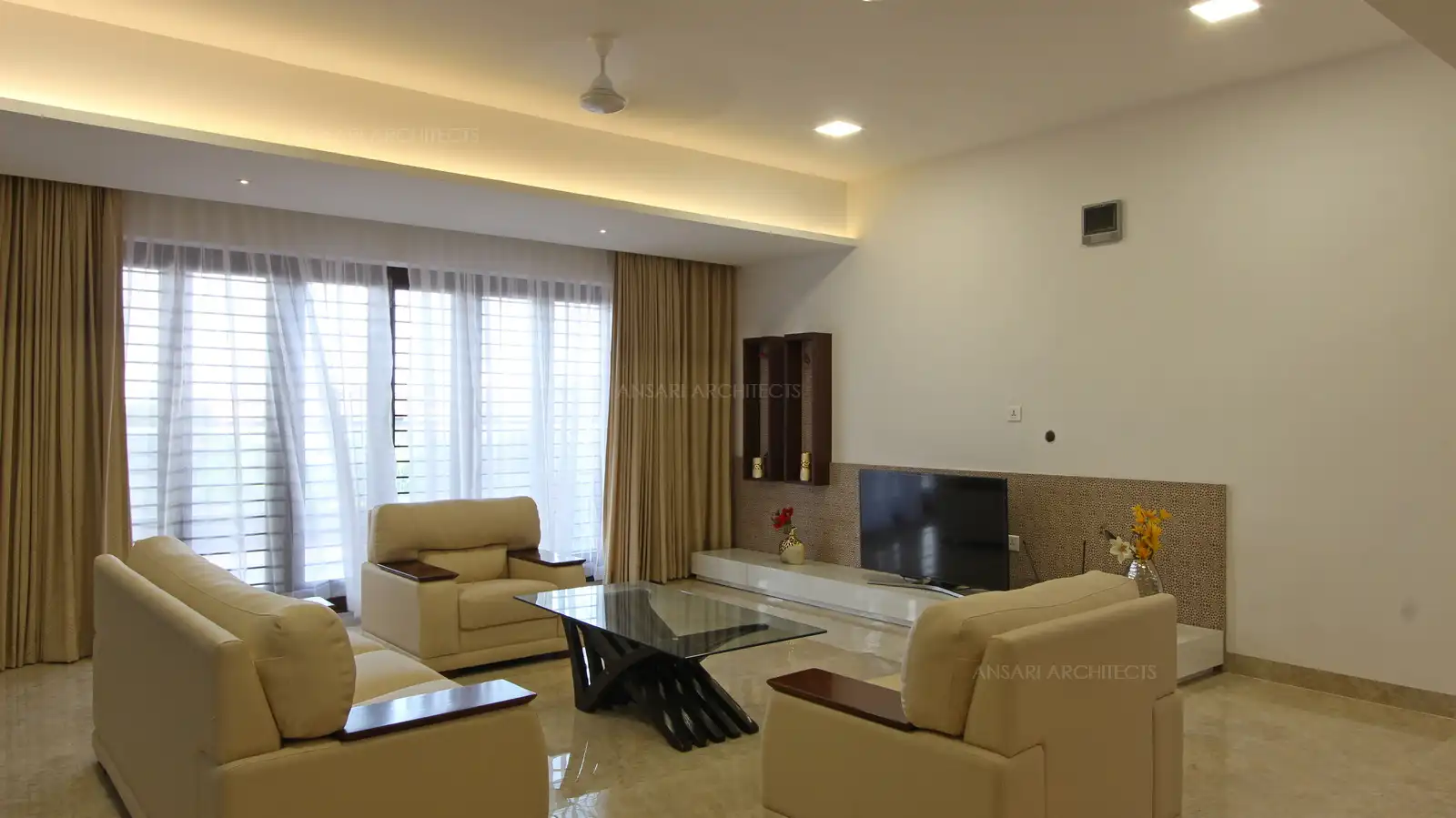 The room is wrapped around with tall glass windows that fill the room with natural light, Curtains maintain a clean look, TV unit here balances the contrasr with the simple panel design with wall niches inviting warm shades.