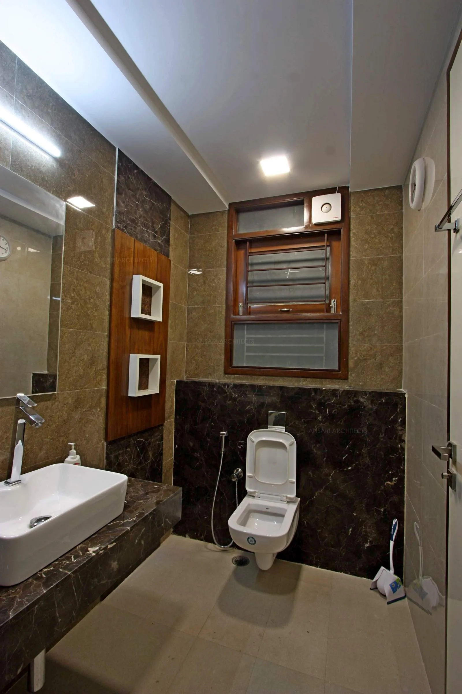 Luxurious bathroom space with wooden cladded floating shelf units, granite top counter for the vanity area with lights along with vitrified tiles dark toned to create a neutral effect.