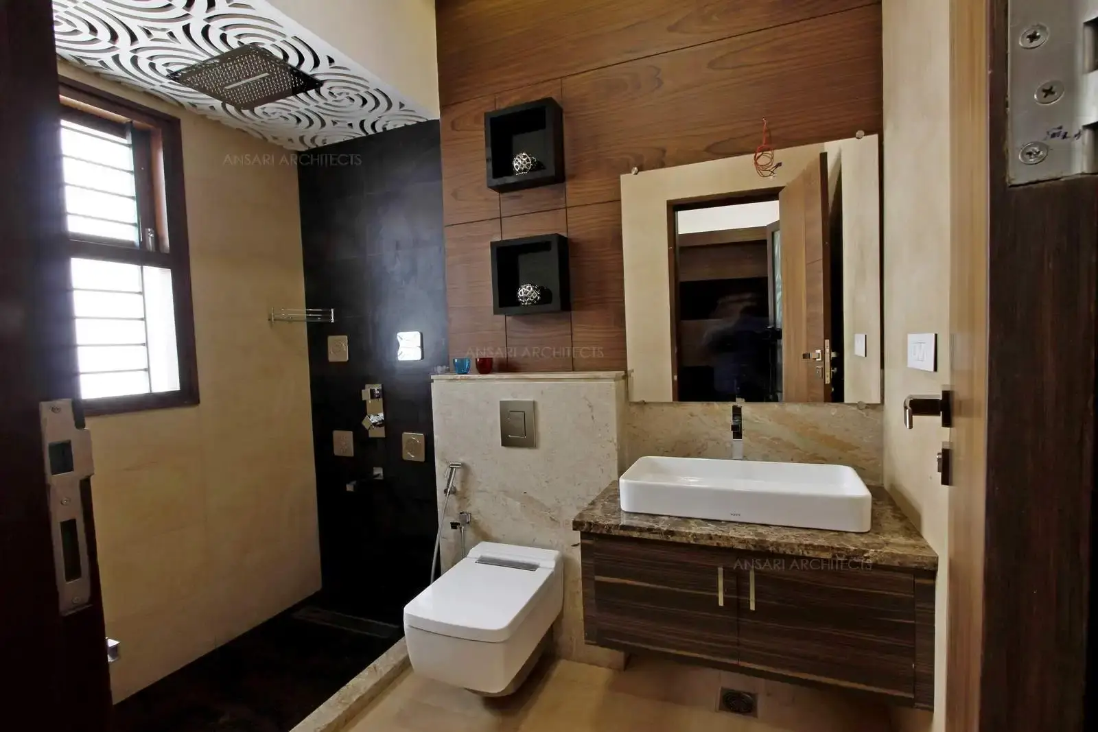 The arrangement of vanity frames the focal point. The elements like wood panelling, marble top washbasin magnify the sense of grandeur. Shower area is sunk to separate wet areas to ensure functionality. Niches are centre of attraction with neutral colour. The space is well lit with natural light.
