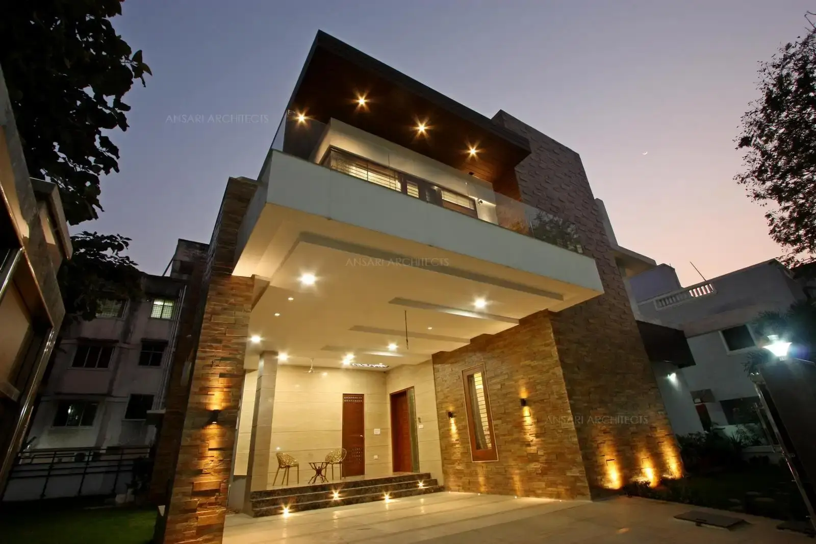 The stone texture brings warmth along with glass, the combination of light styles form an interesting exterior lighting layer. Two-way lighting wall fixtures, Recessed spot lights on the ceiling that subtly highlights different sections of the building. The porch area is welcomes with classic porch furnitures.