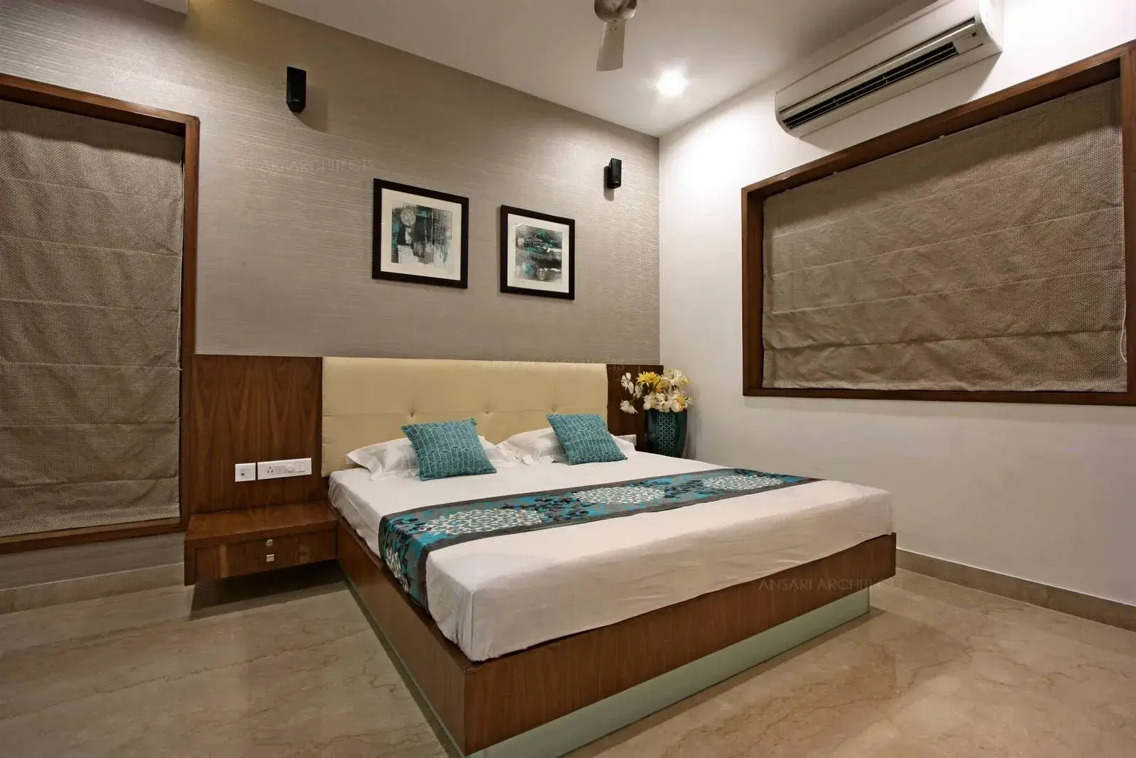 The large fixed glass and window provides natural light for the bedroom and functional side tables are provided for the bed with warm veneer look on the headboard. Wall paper highlights the background.