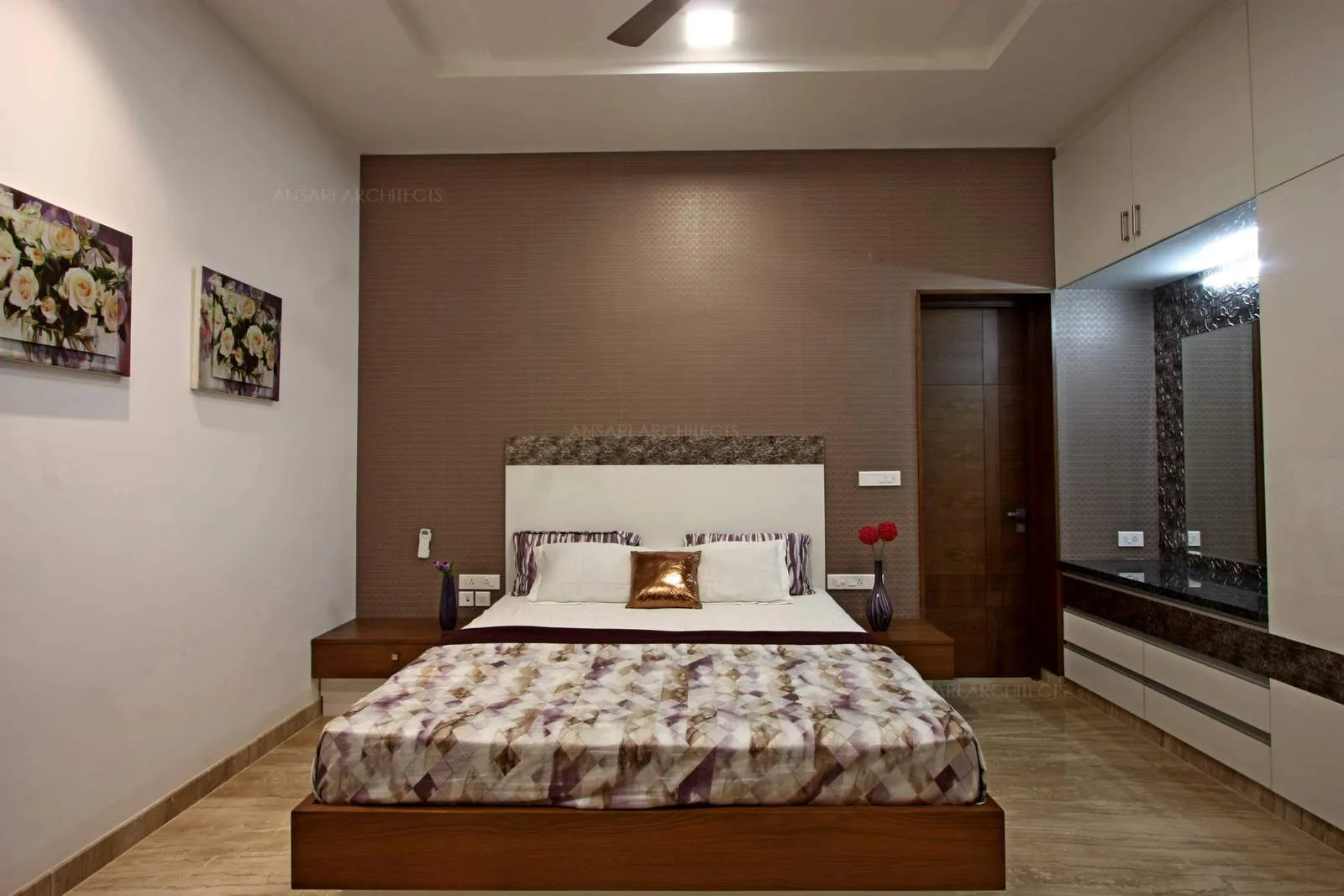 The bedroom in dark brown and white tones. The choice of artwork with floral effects bind the space, wardrobe forms an alcove where vanity unit is situated and camouflages with the walls.