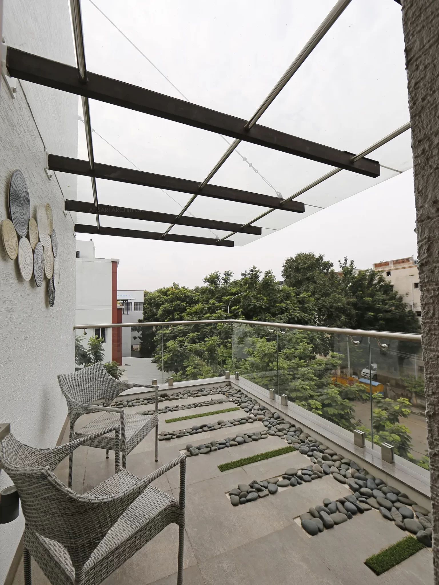 A small deep sitout space with sturdy outdoor seatings and glass canopy shelter which dominates the space with textured walls and hardscaped flooring.