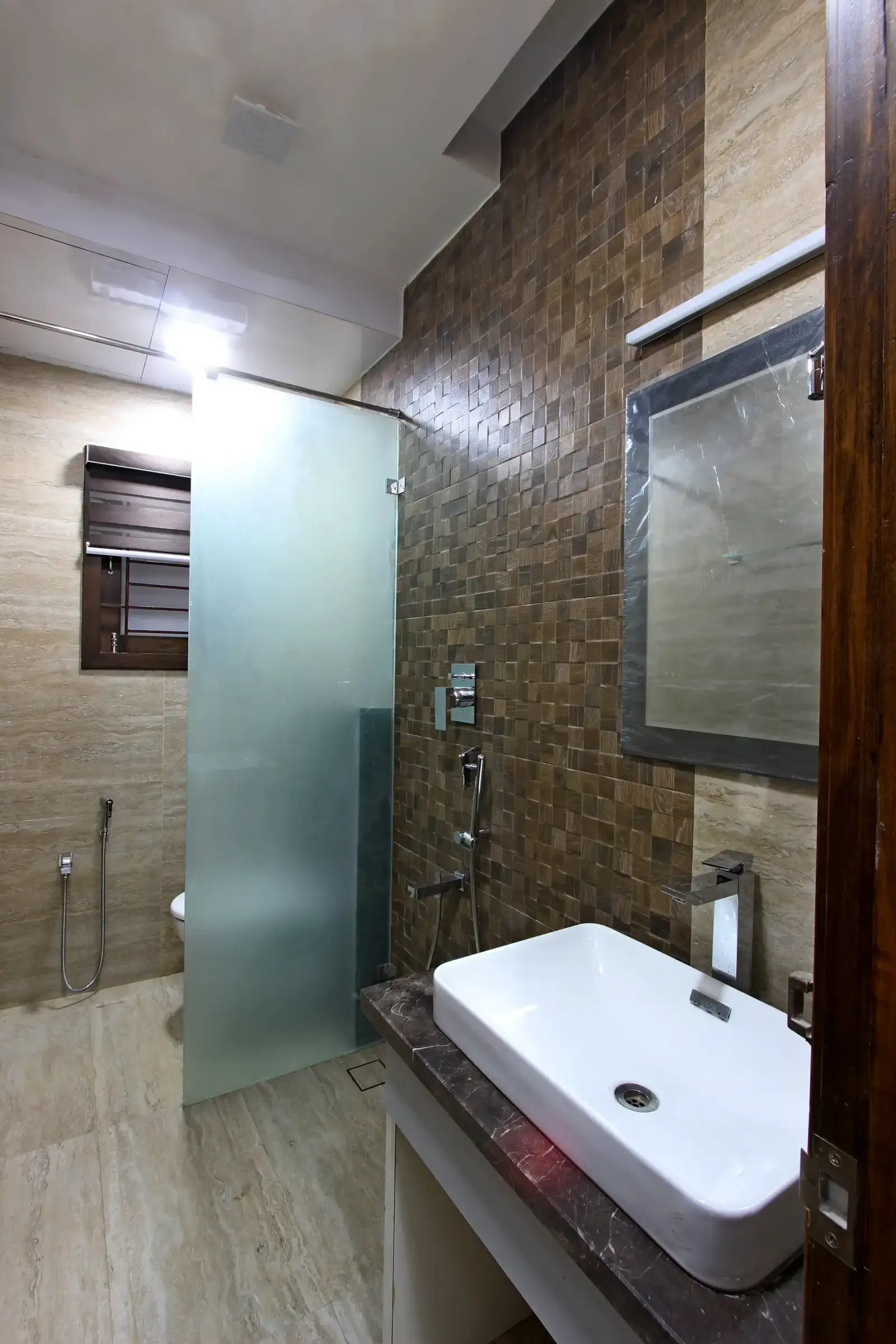 Vitrified tiles with brown outook in walls and glass partition for the shower area and wall mounted lights is highlighted adds elegance to the bathroom space