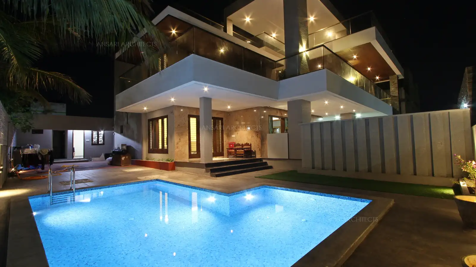 The swimming pool is the focal point. The underlit pool gives a clear view of the floor tiles with the backdrop of rural greens. The L-shaped pool offers a relaxed vibe. The spotlights on walls highlight the textures in compound wall and provides depth.