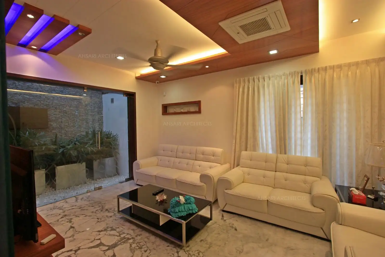 The drawing room in the ground floor looks open and spacious with its white colour scheme combined with wooden finishes on the false ceiling.