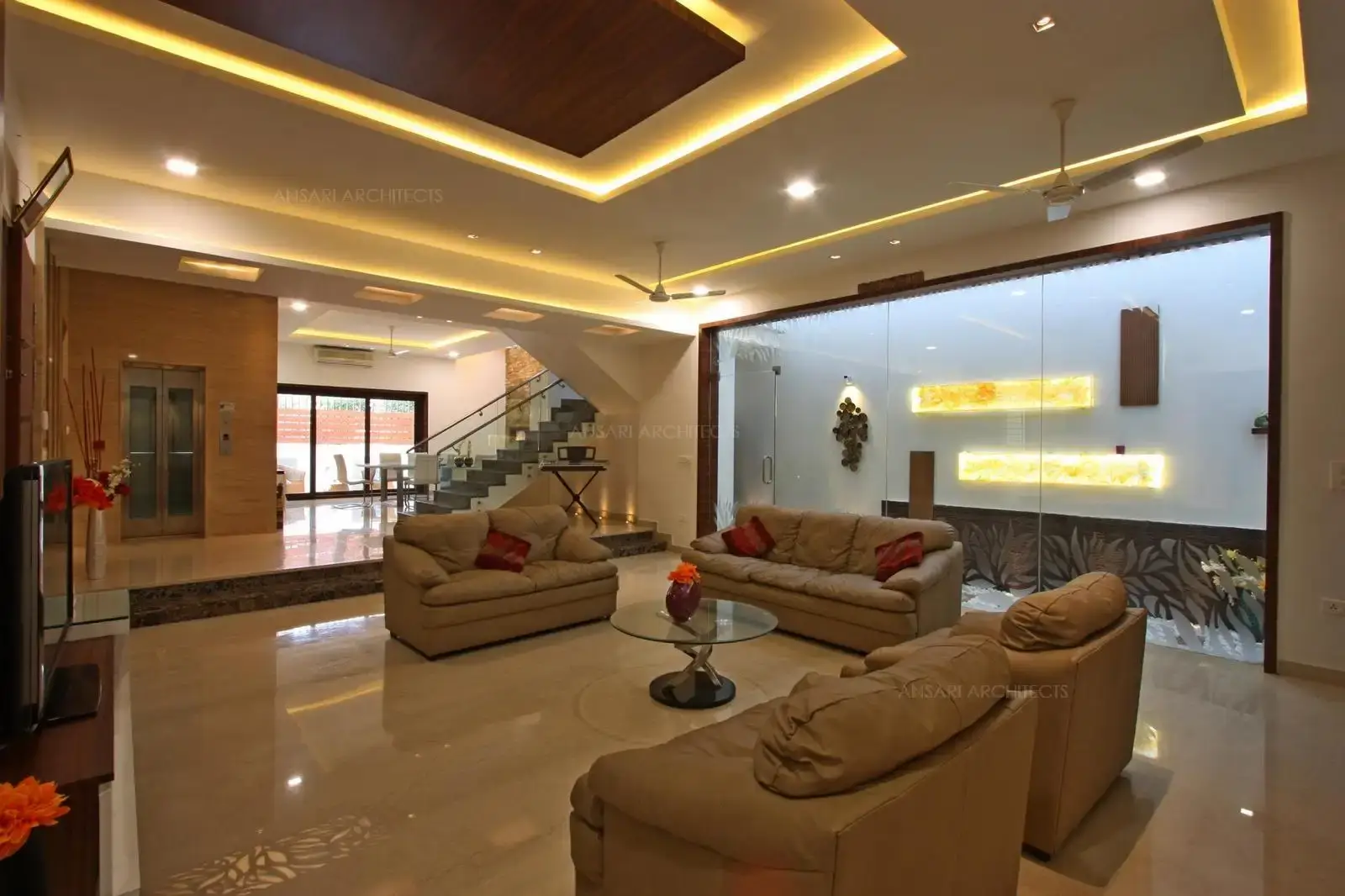 The cove lights enhance the space with marble flooring which creates a contrast with the white walls. The glass top centre table is simple and the entire space overlooks the courtyard which welcomes natural light and extends the visual line the white colour scheme merges with the dark tones of the wood cladding.
