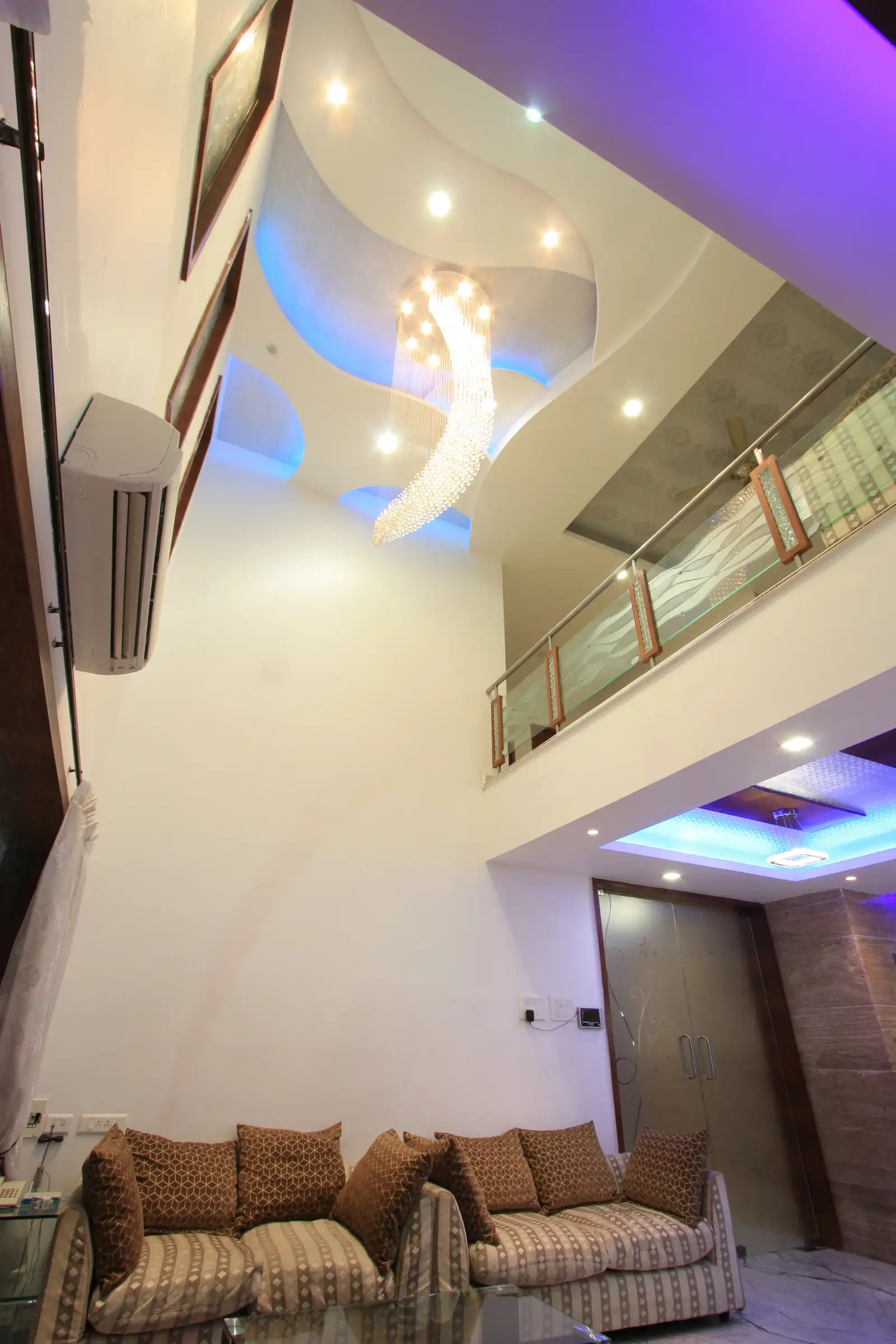 Double height ceiling in the living area has an exclusive chandlier hung on curve design false ceiling with coloured conceal lights.