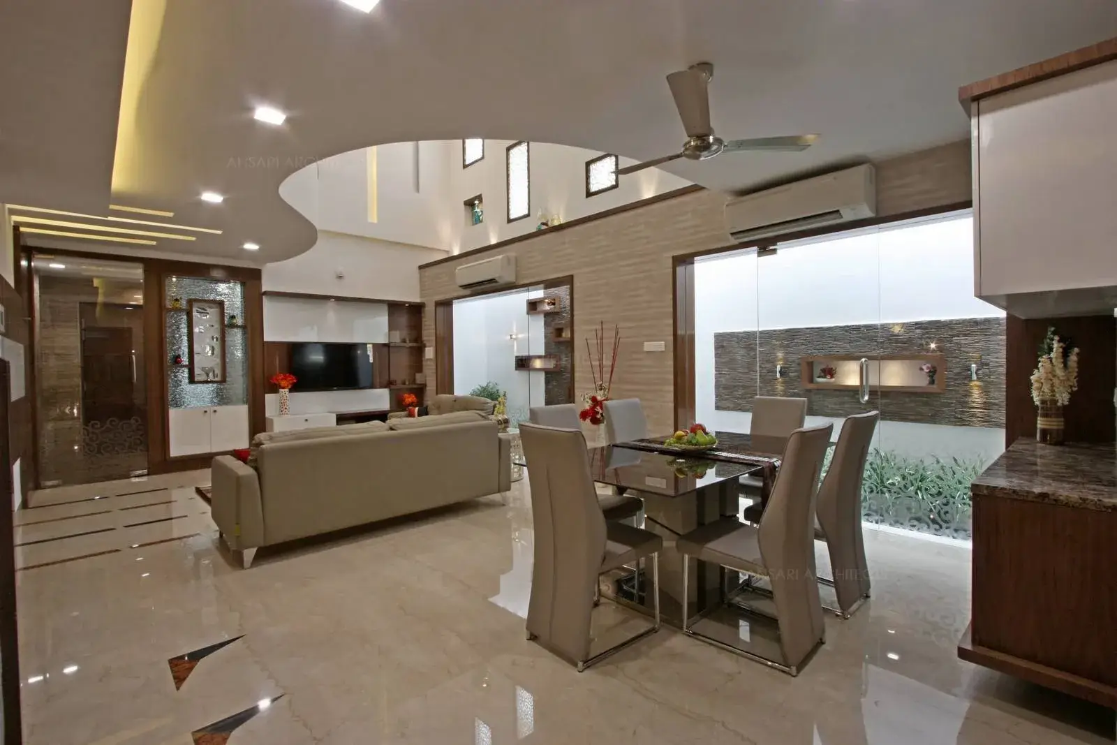 A view of the dining area overlooking the courtyard. The entire space is lit by natural light and glass furnishings complement the space with spot lights and strip lights which highlight the ceiling. Display units and crockery units with glass topped dining table create a contrast.
