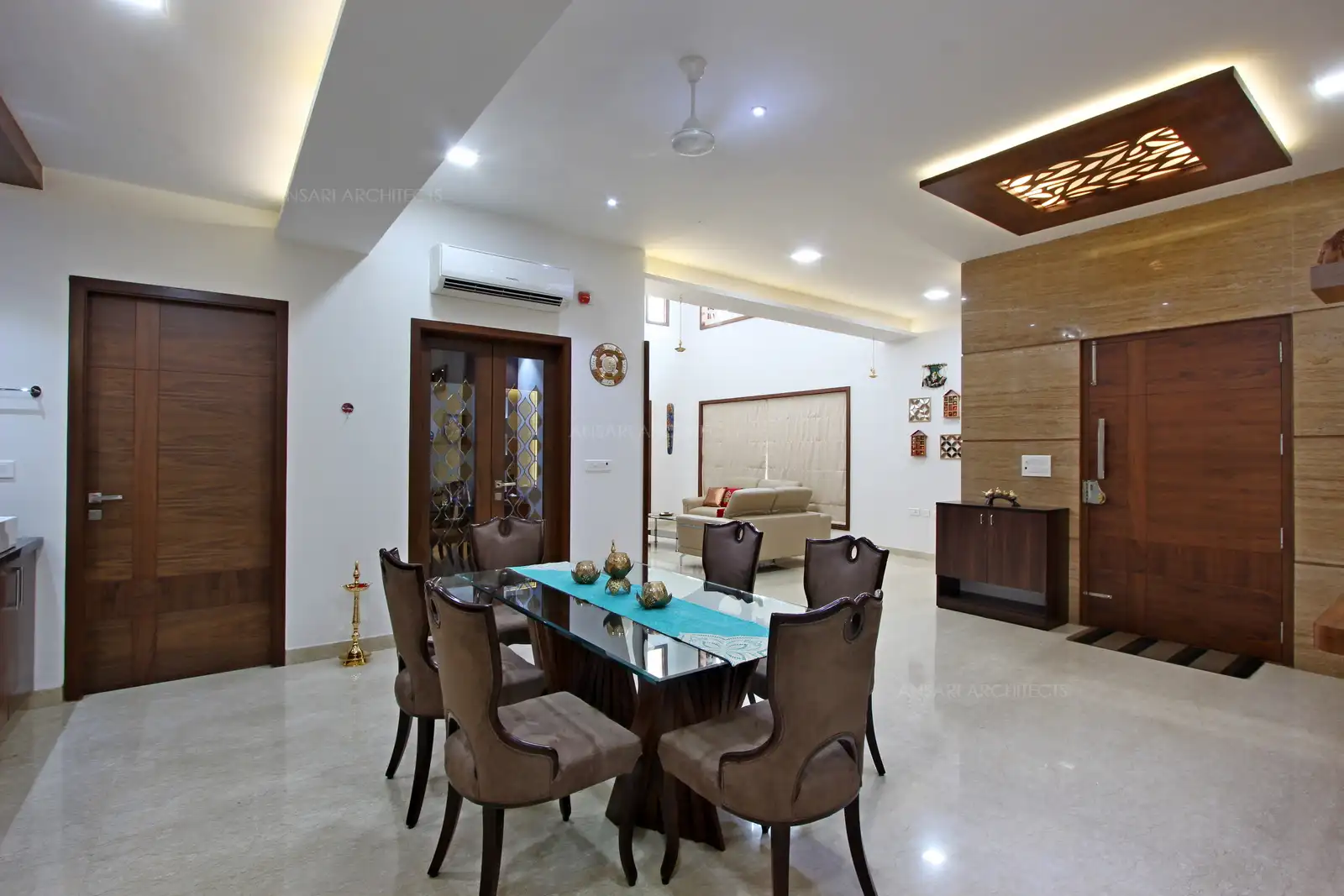 The dining space is classic and elegant with white walls. Glass top table and matching classic cushioned chairs, modern touch of wooden wall unit and solid wooden doors. Light installation provides a eclectic appeal.