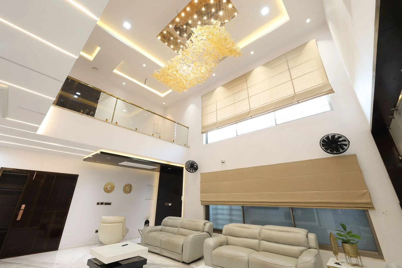 Modern living room with high ceiling, elegant chandelier, beige sofas, and large windows with natural light. Contemporary design and neutral colors create a spacious, cozy atmosphere.