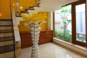 Modern indoor stairway with beige marble steps and a metallic handrail, featuring a tall, shiny silver vase filled with yellow and white flowers, next to a large window showing green plants outside against a yellow and white wall backdrop.