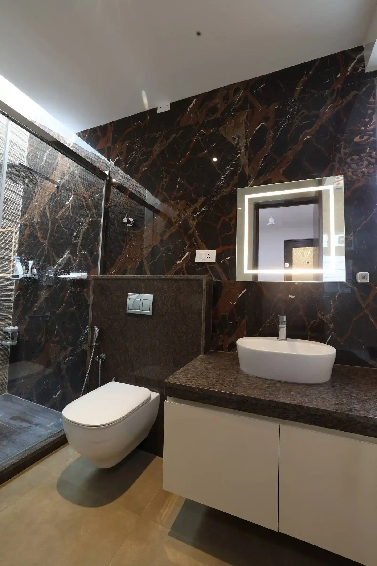 Bathroom features dark marble walls, a glass-enclosed shower, and a sleek countertop with an oval sink and illuminated mirror, combining elegance and functionality.