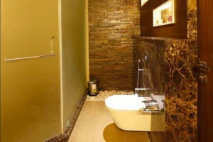 Modern bathroom with a white wall-mounted toilet, brown marble walls, pebble stones along the back wall, metallic trash bin, and frosted glass door with a towel bar.